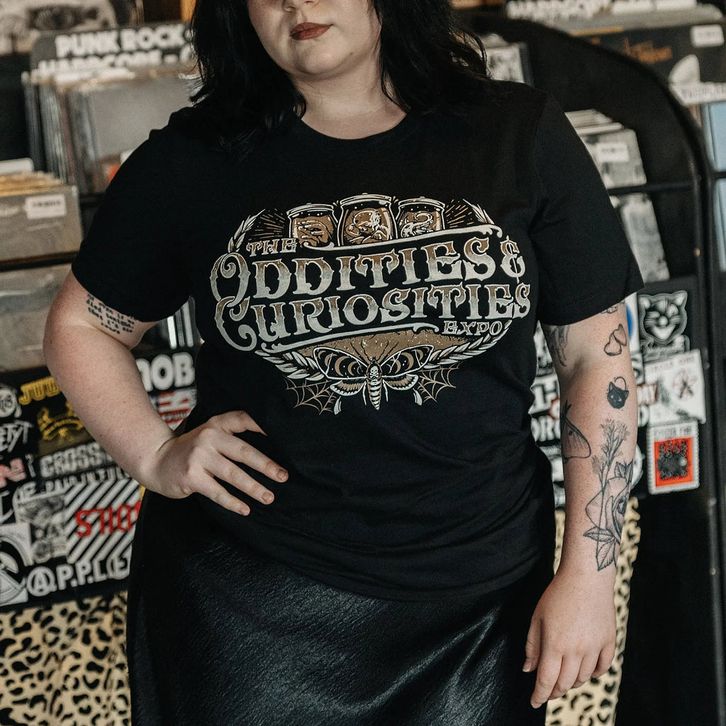 A person wearing a The Oddities & Curiosities Expo - Death Head Moth T-shirt. The large distressed white and beige print reads 'The Oddities & Curiosities Expo' over a moth illustration, photographed in a record-store-like setting with shelves and stickers in the background.