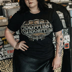 A person wearing a The Oddities & Curiosities Expo - Death Head Moth T-shirt. The large distressed white and beige print reads 'The Oddities & Curiosities Expo' over a moth illustration, photographed in a record-store-like setting with shelves and stickers in the background.