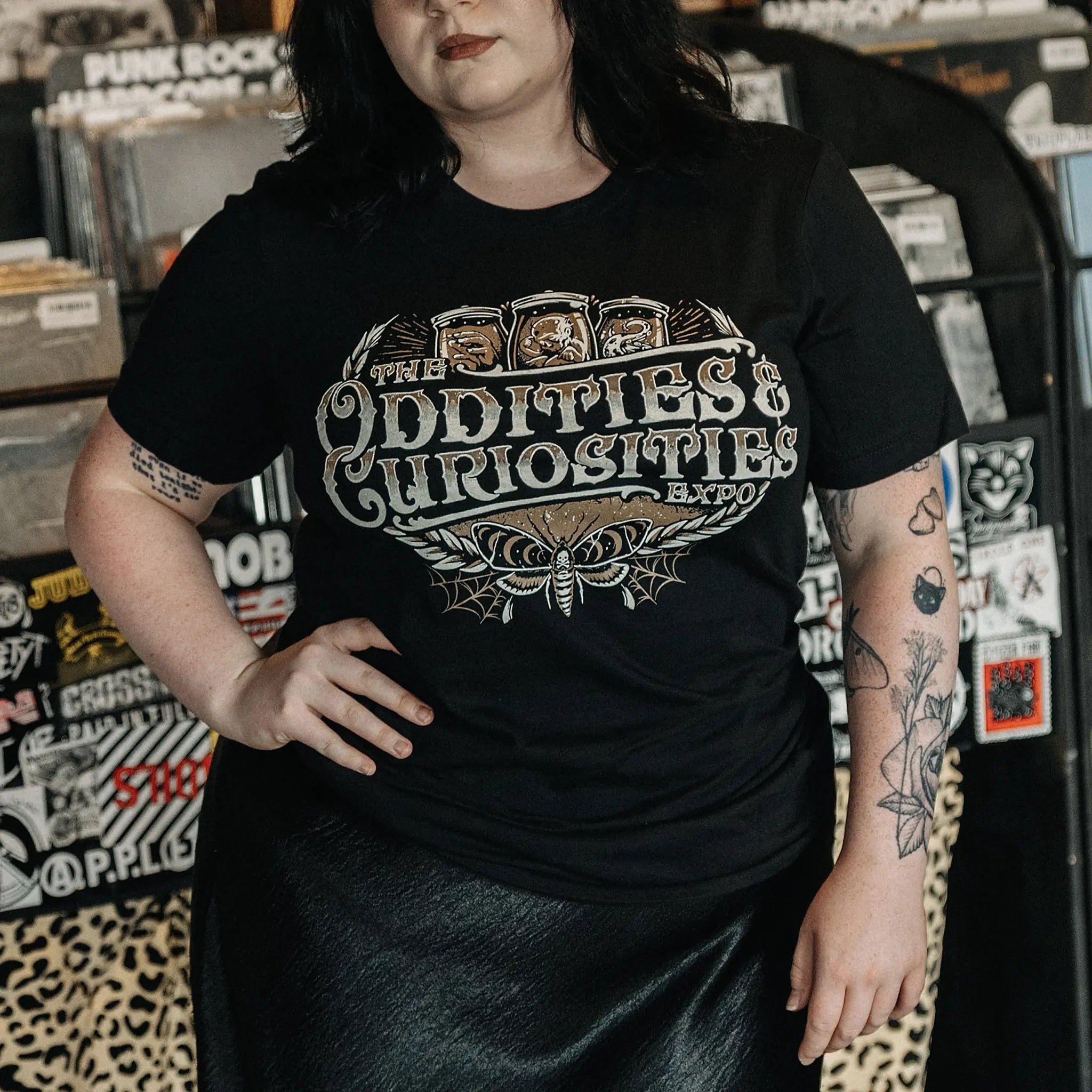 A person wearing a The Oddities & Curiosities Expo - Death Head Moth T-shirt. The large distressed white and beige print reads 'The Oddities & Curiosities Expo' over a moth illustration, photographed in a record-store-like setting with shelves and stickers in the background.
