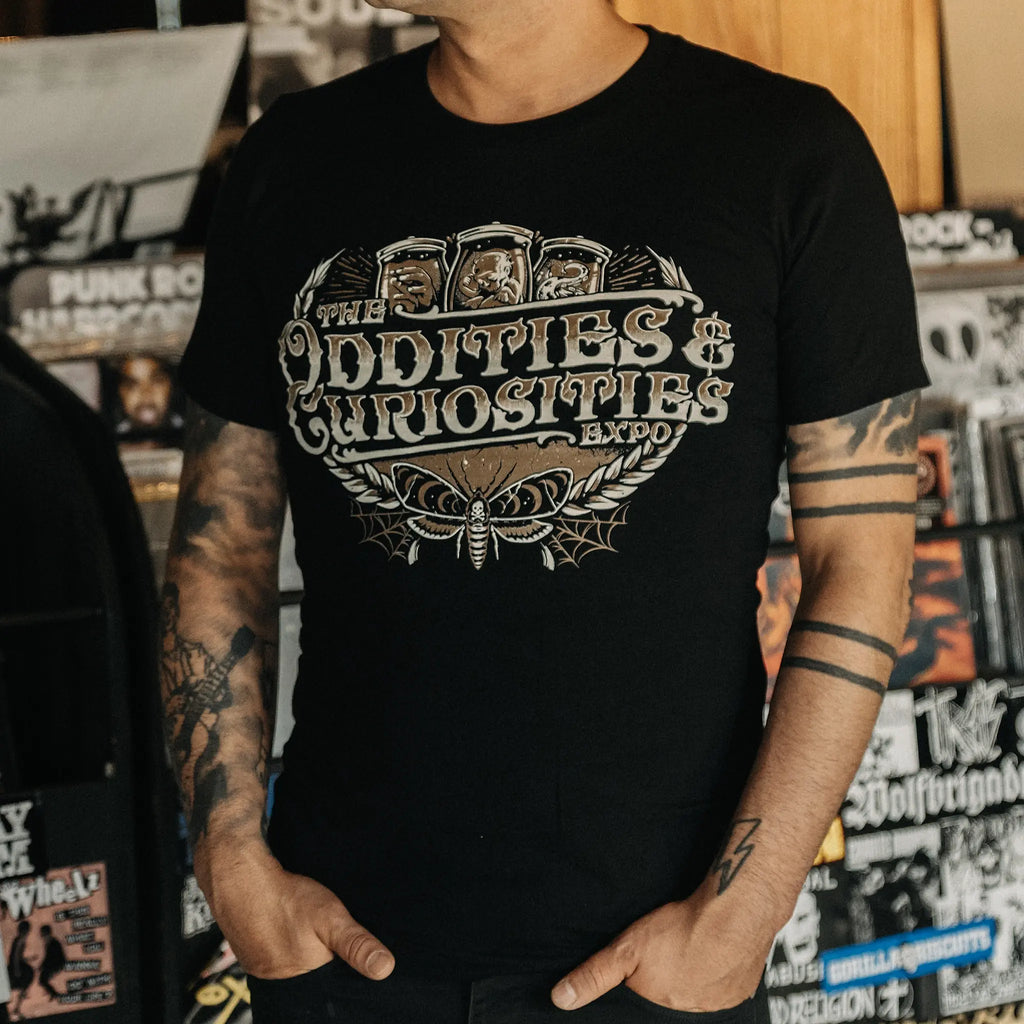 A person wearing a The Oddities & Curiosities Expo - Death Head Moth T-shirt. The large distressed white and beige print reads 'The Oddities & Curiosities Expo' over a moth illustration, photographed in a record-store-like setting with shelves and stickers in the background.