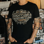A person wearing a The Oddities & Curiosities Expo - Death Head Moth T-shirt. The large distressed white and beige print reads 'The Oddities & Curiosities Expo' over a moth illustration, photographed in a record-store-like setting with shelves and stickers in the background.