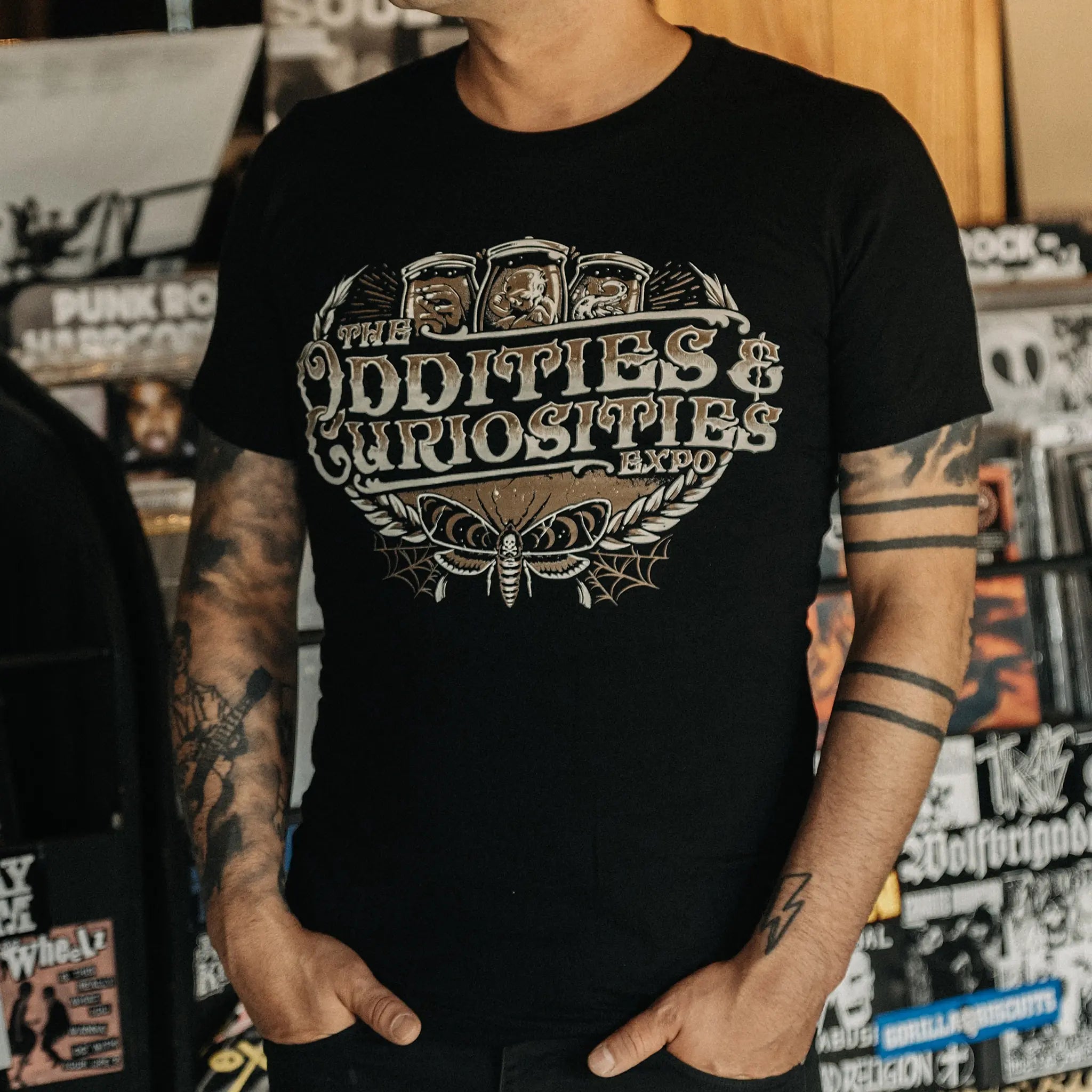 A person wearing a The Oddities & Curiosities Expo - Death Head Moth T-shirt. The large distressed white and beige print reads 'The Oddities & Curiosities Expo' over a moth illustration, photographed in a record-store-like setting with shelves and stickers in the background.